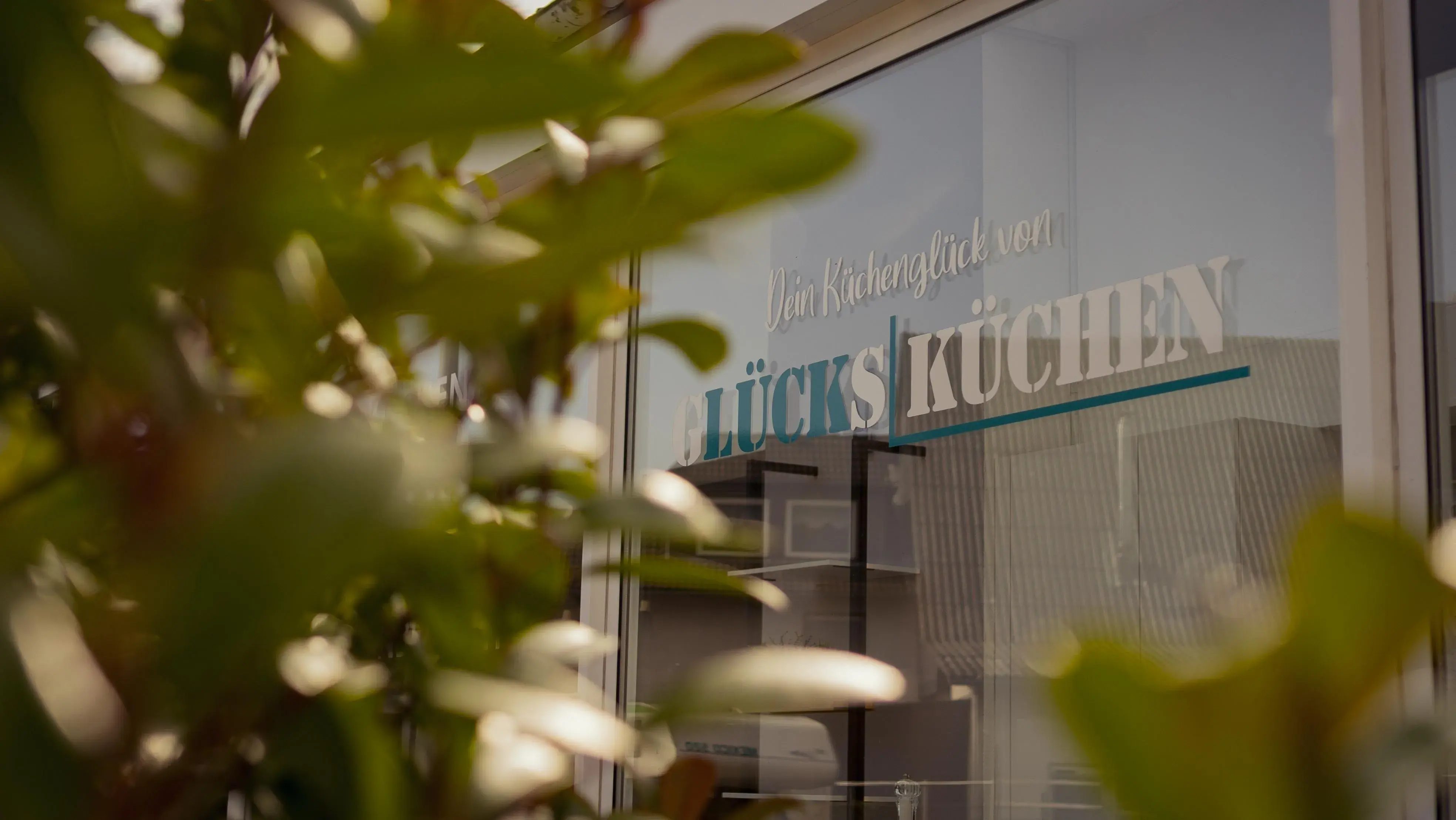 Fenster mit Schriftzug „Glücksküchen“, vorne unscharfe grüne Pflanzenblätter.